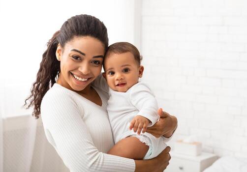 alegre negro joven mamá participación y abrazando adorable bebé chico disfrutando maternidad y niño cuidado en pie en moderno dormitorio a hogar, sonriente a cámara. de la madre día concepto foto