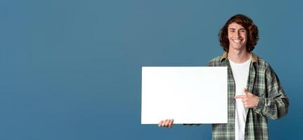 Joyful millennial guy pointing at blank poster on turquoise background, space for your design photo