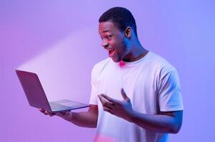 Online Offer. Surprised African American Man Holding Laptop Looking At Glowing Screen, Excited Black Guy Playing Games Or Browsing Internet On Computer, Stranding In Neon Light Over Purple Background photo