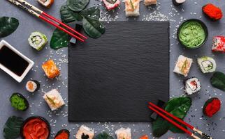 Top view on traditional japanese empty square black slate plate framed with sushi, ginger, soy, wasabi bowls, green leaves and chopsticks on rustic grey backround copy space photo