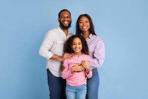 Love And Care Concept. Portrait of African American mum and dad embracing smiling daughter standing isolated on blue studio background. Cheerful man and woman posing with girl and looking at camera photo