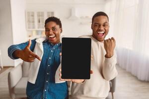Excited Young Black Couple Showing Tablet Empty Screen, Woman Pointing, Man Shaking Fist,Celebrating Good News Standing At Home, Smiling To Camera. Great Mobile Application. Mockup, Blank Space photo