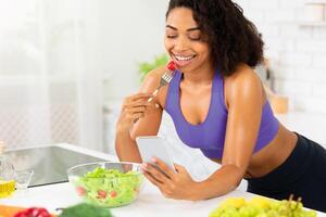 Young african woman eating salad at hone in kitchen, leaning on table and texting on smartphone, using an application photo