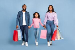 Family Shopping Time. Happy black man, woman and their daughter walking together with colorful shopping bags, holding hands and looking at camera isolated over blue background in studio photo