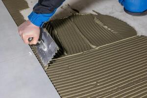 Tiler worker placing or tiling gray ceramic tile in the position over adhesive glue with lash tile leveling system, renovation or recontruction, concept of building photo