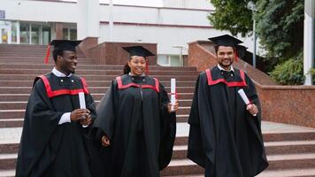 Three multiracial college graduates in graduation mantles walking along university campus and talking video