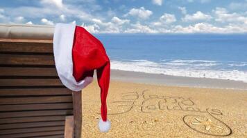 Happy Holidays from the Beach 4K features a beach chair with a Santa hat hanging and gently blowing in the wind with an ocean in the background and Happy Holidays written in the sand video