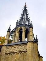 An imposing Gothic-style chapel with a tall, intricately detailed spire reaching into the overcast sky. The structure stands with its arched entrance and stone facade, embodying historical photo