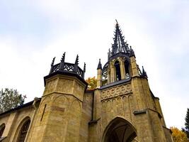 An imposing Gothic-style chapel with a tall, intricately detailed spire reaching into the overcast sky. The structure stands with its arched entrance and stone facade, embodying historical photo