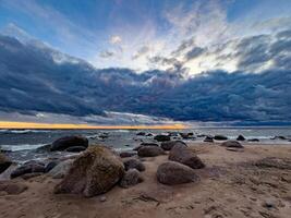 un asombroso ver de un rocoso playa a atardecer, con olas estrellarse en contra el piedras debajo un dramático cielo. oscuro nubes tramo a través de el horizonte, mientras el ajuste Dom yesos un calentar dorado resplandor foto