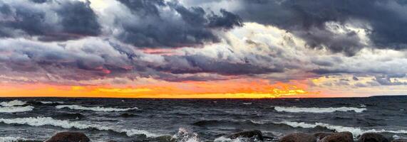 un asombroso ver de un rocoso playa a atardecer, con olas estrellarse en contra el piedras debajo un dramático cielo. oscuro nubes tramo a través de el horizonte, mientras el ajuste Dom yesos un calentar dorado resplandor foto