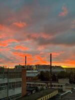 A captivating view of an industrial cityscape during a fiery sunset, where the sky is illuminated in intense shades of red and orange. The chimneys and rooftops create a sharp contrast against the photo