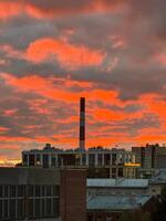 A captivating view of an industrial cityscape during a fiery sunset, where the sky is illuminated in intense shades of red and orange. The chimneys and rooftops create a sharp contrast against the photo