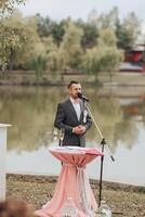 A man stands at a podium in front of a lake, singing into a microphone photo