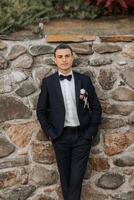 A man in a suit and bow tie stands in front of a stone wall photo