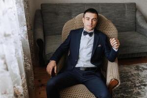 A man in a suit and bow tie sits in a chair photo