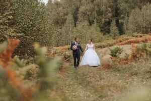 A bride and groom are walking through a forest, holding hands and smiling photo