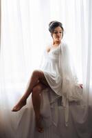 A woman in a white dress sits in front of a window photo