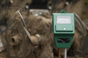 Close-up of a green soil moisture meter in a garden setting with blurred muddy background, photo