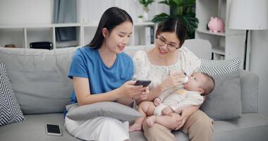 Portrait of Two happy women are enjoying watching something together on mobile phones while bottle-feed the baby boy on sofa in the living room. Happy family time photo