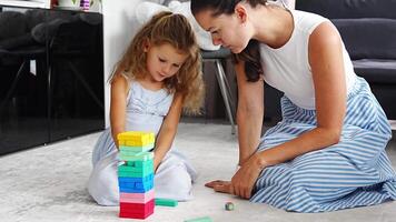 Little girl with her mother playing Jenga board game at home, taking colorful blocks from wooden tower. High quality 4k footage video