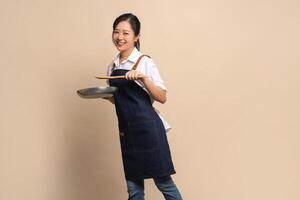 Beautiful and attractive Asian female coffee shop owner wearing apron. She holds a frying pan and a wooden spatula on a beige background. photo