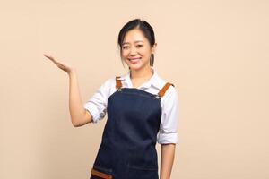 Beautiful Asian woman in apron pointing at the empty space for your ad on beige background photo