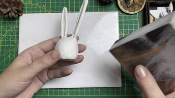 Women's hands polish clay figurines in form of white rabbit with sandpaper. Tools for modeling on desktop. Jewelry designer's workshop. Handmade and craft. video