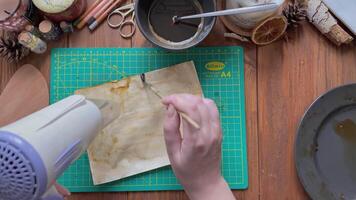 designer åldrar scrapbooking papper och måla den med borsta. konstnär dekorera papper med vatten och kaffe och torr den med hårtork. penna, sax på trä- tabell. fritid i karantän. video