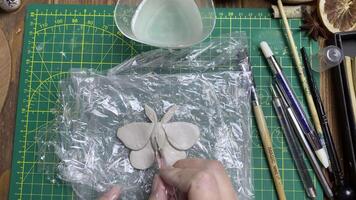 Female's hands make shape for clay moth with metallic stick. Tools for modeling and design of ceramic souvenirs. Hobbies, handmade and crafts. Cup of water and brush on wooden table. video
