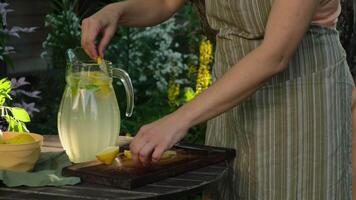 Hands put lemon slices in jug of homemade lemonade. Making summer refreshing lemonade in backyard garden. video