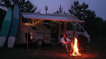 A man is sitting by the campfire in the evening, wrapped in a blanket. Evening rest near a mobile home video