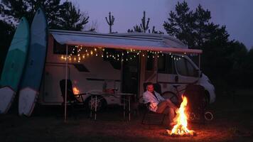 A man is sitting by the campfire in the evening, wrapped in a blanket. Evening rest near a mobile home video