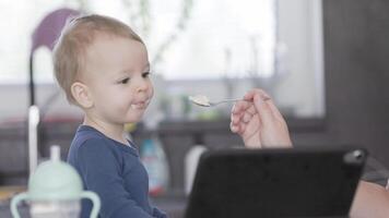 Mother feeds her baby boy with a spoon as he makes funny faces and watching watching a cartoon fairy tale on a tablet video