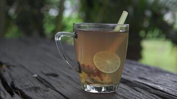 A glass of tea with a slice of lime and lemon grass in it sits on a wooden table. video