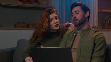 Smiling Caucasian family married couple husband wife man woman making purchase in online store choosing goods on Internet together using laptop in evening at home discussing order delivery arguing video