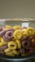 Concept of food and breakfast. Stock footage. Close up of colorful ring corn flakes in a transparent glass bowl on dark table surface and grey wall background. video