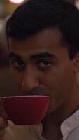 Man drinks tea and winks. Media. Handsome man drinks coffee and winks on background evening lights. Man flirts over cup of coffee in outdoor at cafe in evening video