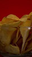 Close up of potato chips inside the transparent glass bowl. Stock footage. Crispy junk product in a small deep dish lighted isolated on red background. video