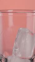 Close up of ice cubes falling into a cocktail empty glass isolated on pink background. Action. Making a cocktail with ice. video