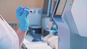 Laboratory worker holds a vial and mixes it by hand in hospital. Technician woman works with blood samples while standing close to the automated machine in clinic. Close-up. video