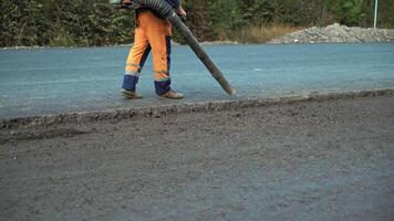 Worker in protective clothes blows out the pieces of old asphalt from the pits at road construction site. Road works concept. Worker is using compressed air to blow off dust on the road. video