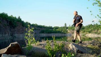 Bald man in t-shirt, shorts and trainers walking with backpack on the big stones near beautiful lake. Natural background of scenic canyon with a clear lake in summer. video