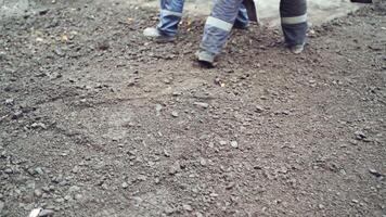 Legs of workers in protective uniform and boots with shovels leveling up the road. Team of workers preparing the ground for making new asphalt on a street. Close-up video