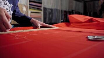 Woman tailor is working at table with red cloth in atelier. Skilled seamstress is drawing sketch lines with a soap or a chalk and a ruler on red fabric. video