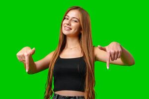 Young Woman in Black Tank Top Points Downward With Both Hands Against Green Screen photo