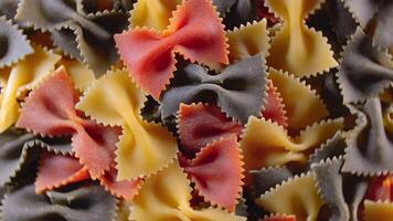 Uncooked Vibrant Colored Farfalle Pasta. A Culinary Canvas of Multicolored Bow-Tie Macaroni, Textured Background - Top View, Angular Rotation. Turning Colored Dry Pasta. Raw Macaroni - Rotating video