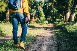 unrecognizable man with backpack and boots holding a camera in front of a path in a forest, nature travel and hiking concept. photo