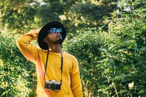 Young Latin American man in a forest wears a yellow sweater with a camera and a hat, looking around. travel and natural recreation concept. photo