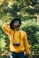 Young Latin American man in a forest wears a yellow sweater with a camera and a hat, looking around. travel and natural recreation concept. photo